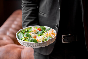 A person in a suit holds a stylish plate of fresh salad filled with greens, shredded vegetables, and cherry tomatoes, set against a cozy background with leather seating.