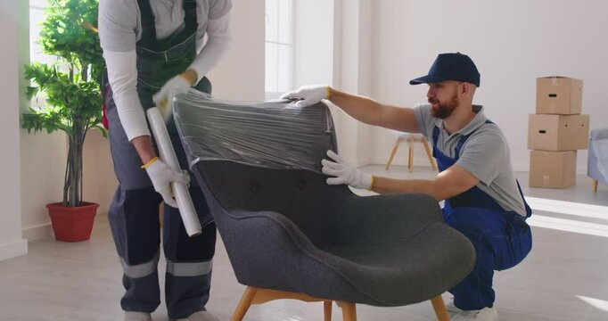 Portrait of a two young smiling male movers packing furniture in the living room. Moving service men workers wrapping chair with plastic wrap. Move, moving day and relocation concept. 4k video.