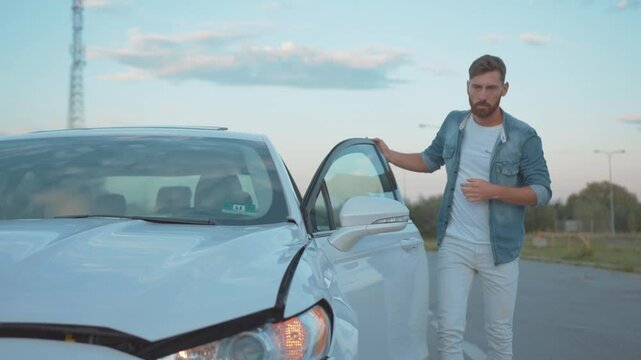 Upset man driver calling car services looking at his crashed vehicle after accident staying in city outskirts at sunset.