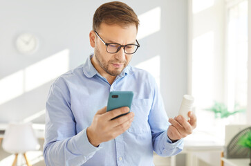 Portrait of young confident man standing in living room at home with a plastic bottle of medical pills browsing internet on mobile phone reading drug use instruction or buying medication online.