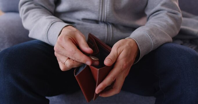 Close up cropped view of poor upset man sitting on sofa and opening wallet with small amount of coins. Displeased person with no money having financial problems, lacking budget, bankruptcy or debts.