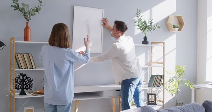 Happy husband, wife doing creativity decor, hanging frame picture on wall, moving settling together in new house, couple decorating own home relocating, ownership, design with love, family relocation