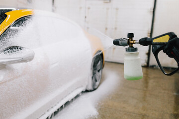 Application of soapy foam with a water gun for sports car cleaning at the service. Worker sprays soap solution with pressure from water jet for detailing car wash.