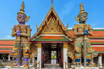 Two giant Yaksha guardian statues stand majestically at the entrance of the magnificent Wat Phra Kaew, adorned with intricate golden details and a striking multi-colored roof beneath a clear blue sky.