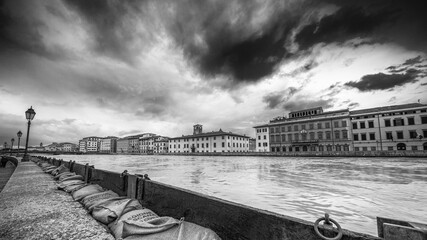 Obraz premium Dramatic view of the swollen Arno River at sunset with flood defenses along Pisa’s Lungarni