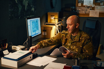Military officer in camouflage uniform reviewing documents at office desk with computer monitor and...