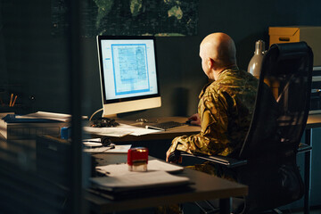 Person dressed in military uniform sitting at office desk facing computer screen with map and...
