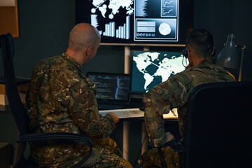 Military personnel evaluating strategies using computers and charts in mission control room. Engaging in critical analysis with global maps and data displaying on screens