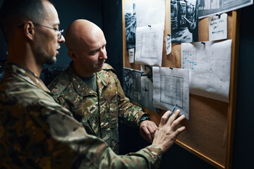 Two military personnel examining and discussing strategic plans on bulletin board inside a dimly lit room, each pointing at different sections on the papers during the briefing
