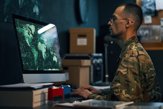 Military personnel working on computer, analyzing global data with maps on screen in an office. Wearing camouflage uniform and focused on task at hand