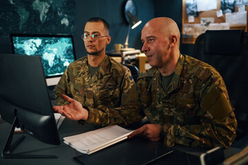Two military personnel in fatigues examining screen and documents in strategy room with maps on wall around. Engaged in detailed strategic planning without distractions © DragonImages