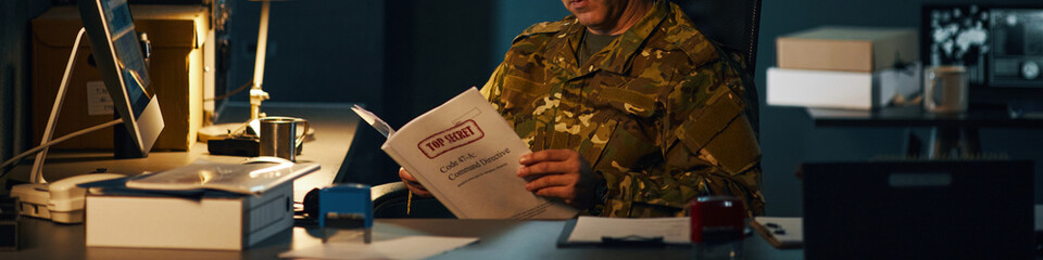 Military personnel dressed in camouflage examining top secret documents in an office setting, surrounded by files and office equipment, dim lighting creating a serious atmosphere © DragonImages