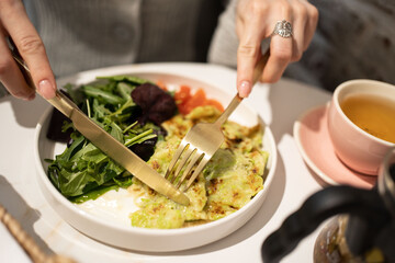Girl in grey at the table with breakfast. Zucchini fritters with salad and tomatoes healthy vegan meal
