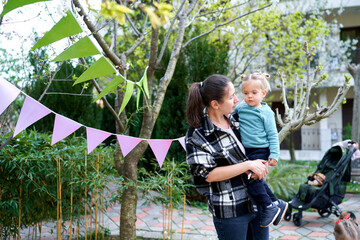 Mom looks at the little girl in her arms standing near a green tree decorated with multi-colored flags