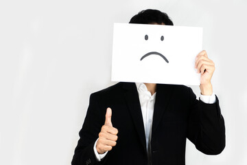Businessman covering face with a sad face drawing and giving a thumbs up, conveying mixed feelings...