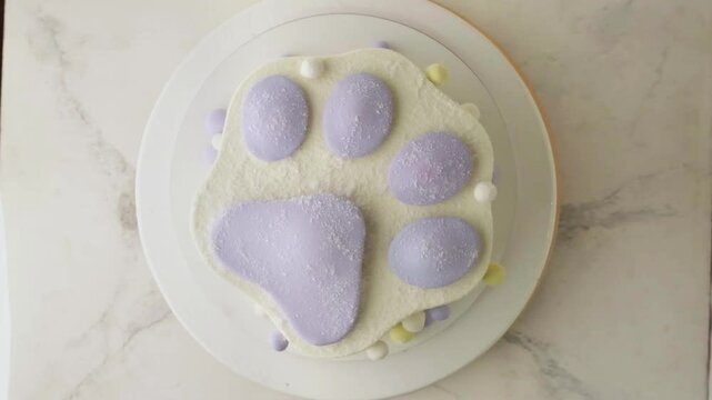 Birthday cake in the shape of trendy squish squishy paw on the white marble background. Flat lay of Taba paw shaped cake for a child with white cream cheese frosting spinning on the cake stand