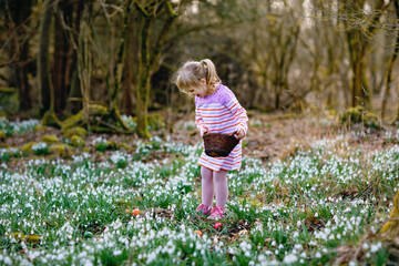 Little girl in pink dress making egg hunt in spring forest on sunny day, outdoors. Cute happy child...