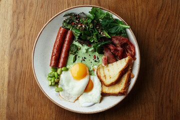 Delicious full breakfast plate with fried eggs, grilled sausages, crispy bacon, toasted bread, fresh green salad, and creamy herb sauce. Healthy and nutritious morning meal on a wooden table