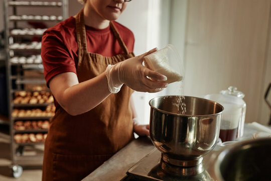 Cropped shot of female baker pouring coconut flakes from measuring cup into stainless steel bowl scaling ingredients for bread dough in bakehouse kitchen, focus on hands in vinyl gloves, copy space