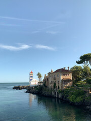 Naklejka premium A scenic view of a coastal area in Cascais, Portugal, with a lighthouse, palm trees, and a historic building by the water, under a clear blue sky.