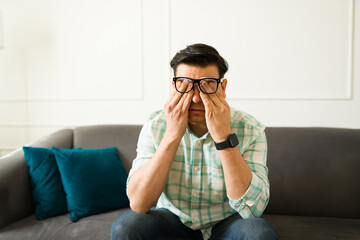 Exhausted latin male rubbing weary eyes, experiencing workplace stress while resting on living room...