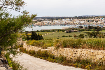 Scenic countryside view of Marsaxlokk, Malta. A beautiful rural road in Malta leading towards Marsaxlokk Bay, featuring traditional stone walls, green fields, and a picturesque fishing village