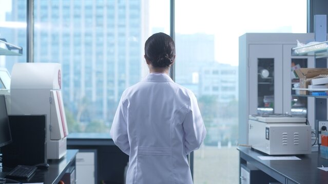Female researcher standing with back to camera in laboratory surrounded by scientific equipment and overlooking cityscape through large window