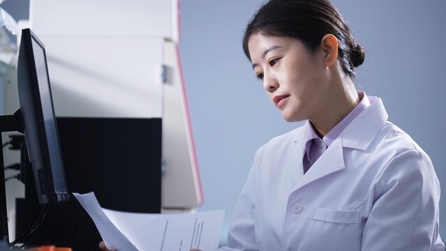 Female doctor in white lab coat focusing on computer screen while reviewing medical documents and data files