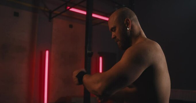 Focused male athlete with no shirt tying a bandage on his wrist while doing strength training in a gym with red neon lights