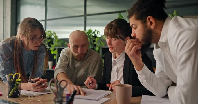 Diverse team of office workers gathers around a table analyzing data on sheets of paper and brainstorming strategies for their next steps