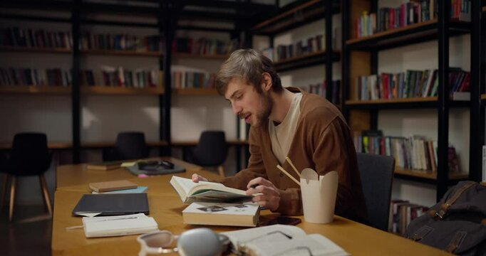 A guy with a mustache and beard sits at a table opens books and takes notes while studying in the library