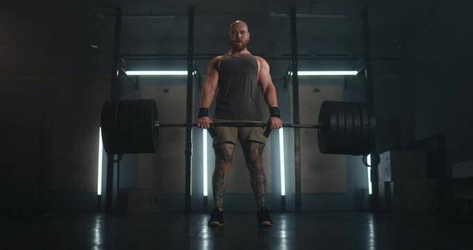 Front view of a large and muscular male athlete lifting heavy weights during a deadlift in a gray gym