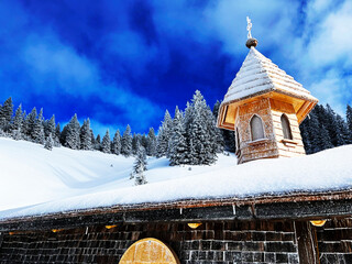 Low angle view of a wooden chapel in the mountains covered in snow against the blue sky with a mountain forest in the background