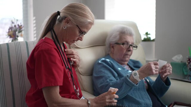 A healthcare provider in red scrubs helps an elderly woman sitting in a comfortable chair. The interaction is warm and supportive in a bright room.