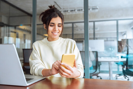 Hispanic business woman manager using cell phone mobile app, laptop. Smiling Latin or Indian student girl holding smartphone sitting in office or modern library, working online on gadget, copy space.