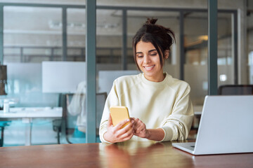 Smiling female indian or latina freelance entrepreneur business girl holding mobile cellphone, using digital technology for work. Middle eastern woman student studying on smartphone laptop pc computer