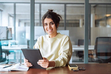 Portrait of middle eastern student or worker using tablet pc financial application standing in office. Smiling indian or latin hispanic business woman holding digital pad computer, looking at camera.
