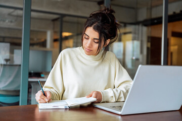 Young latina or indian business woman using laptop pc working at desk writing notes, watching educational online webinar. Female student looking at computer screen device, studying in modern office