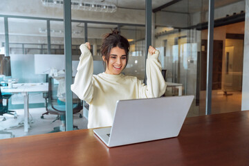 Happy excited young latina hispanic student looking at laptop pc computer doing yes triumph win gesture. Smiling indian business woman successful winner celebrating victory winning achieve in office
