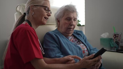 A healthcare worker guides an elderly woman in using her remote at a home setting. The interaction highlights technology support for seniors while sitting comfortably.