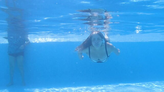 Adult woman swimming underwater in a pool, waving her arms and floating peacefully, blue tiled pool, crystal-clear water, sunny and relaxing vacation day. Active adult lifestyle concept