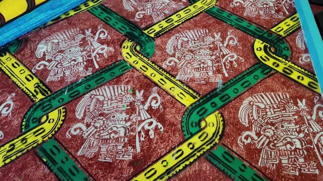 A colorful patterned carpet with a green and yellow design. The carpet is red and white. The design is of a person with a headdress and a snake