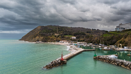Italy, March 15 2025: aerial view from the drone of the Vallugola Bay in Pesaro with its small port, the San Bartolo cliff, the pebble beach at sunset with a threatening storm sky