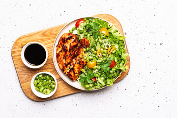 Healthy diet dinner concept. Fresh green salad with edamame beans and cherry tomatoes and chiken breast with teriyaki sauce in the serving board top view.