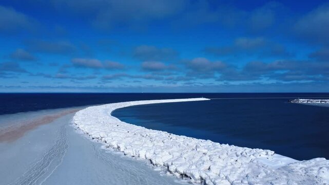An arched wall of white ice clinging to artificial wave-cutting riprap protects the entrance to a harbor from the ocean beneath a panoramic cloudy blue sky that cuts across the horizon.