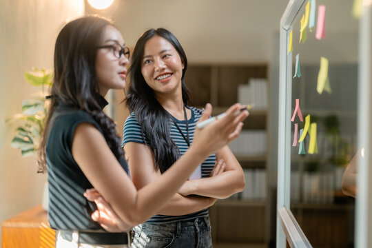 Asian businesswomen discussing and sharing ideas using sticky notes on glass wall in modern office