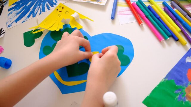 Preparing for Earth Day in the classroom. Top view of table with crafts, child making paper crafts for Earth Day, painting and drawing pictures, creating earth like Planet Earth with smiling face