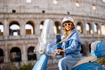A fashionable woman in a blue outfit rides a classic scooter near the Colosseum in Rome, Italy. The...