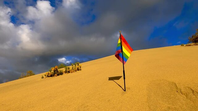 Rainbow flag waving gently in the wind, planted on the top of a sand dune, with some sparse vegetation in the foreground and a blue sky with some clouds in the background, slow motion