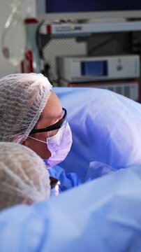 Two doctors wearing white caps and glasses sit near the patient's legs covered with blue sheet. Female surgeons perform gynecologic operation. Vertical video.